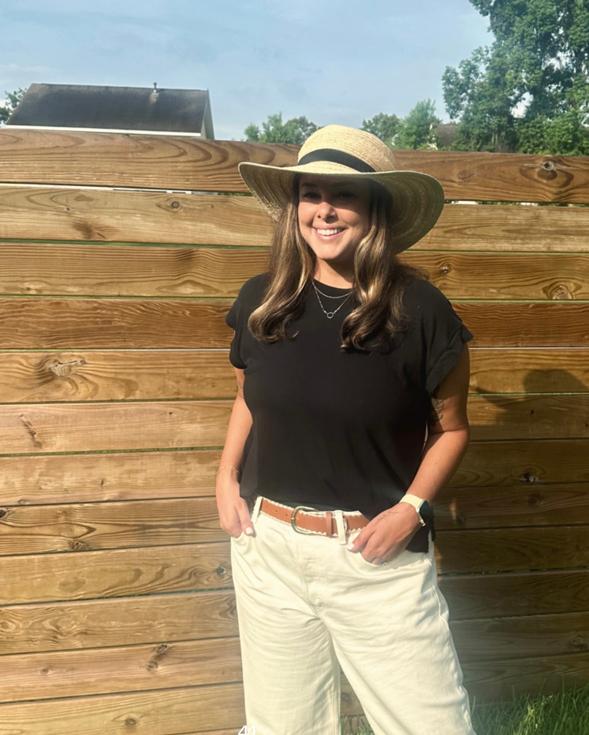 A smiling woman with long brown hair stands outdoors in front of a wooden fence.